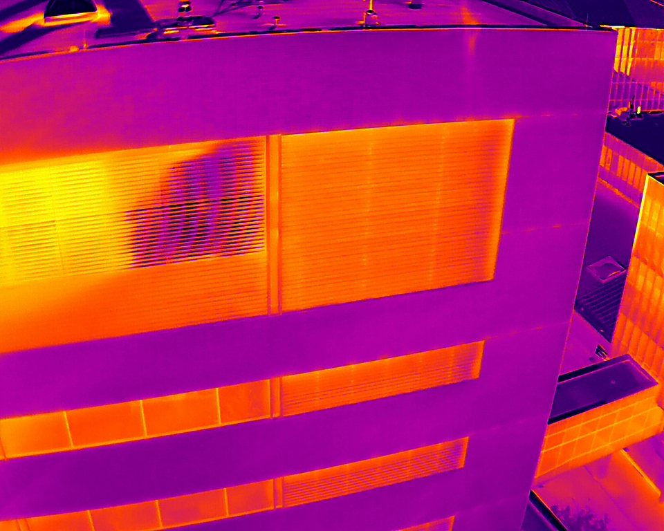 Aerial infrared image of a mid-rise building facade. Horizontal window bands glow bright orange and yellow against cooler magenta spandrel panels, and one window bay mid-frame reads noticeably cooler than the bays beside it.